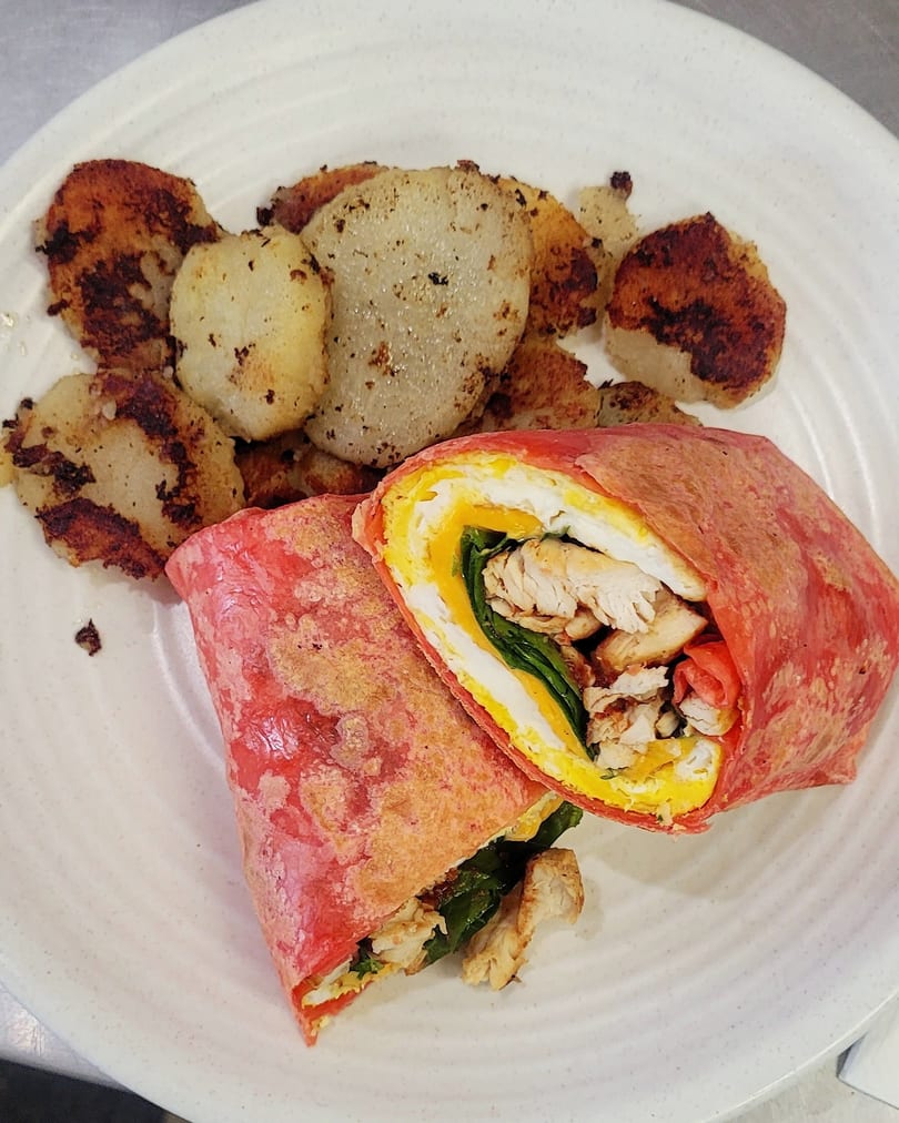 A red tortilla wrap filled with chicken, egg, and spinach, served with roasted potatoes.