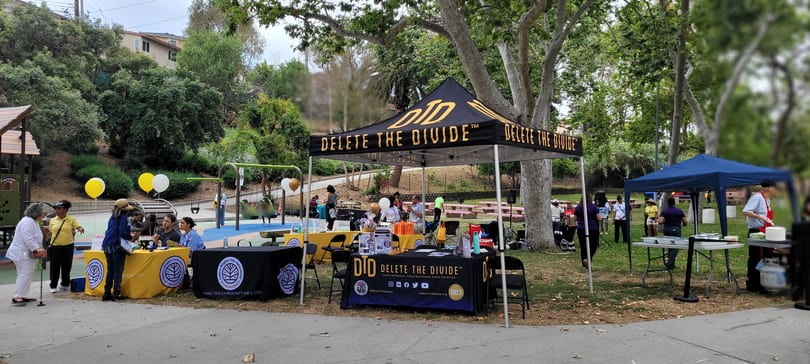 A park event with "Delete The Divide" tents, people, and tables near a playground.
