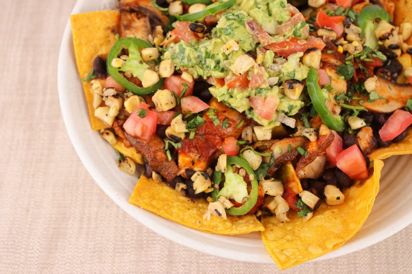 Close-up of loaded nachos with guacamole, tomatoes, corn, black beans, and jalapeños.
