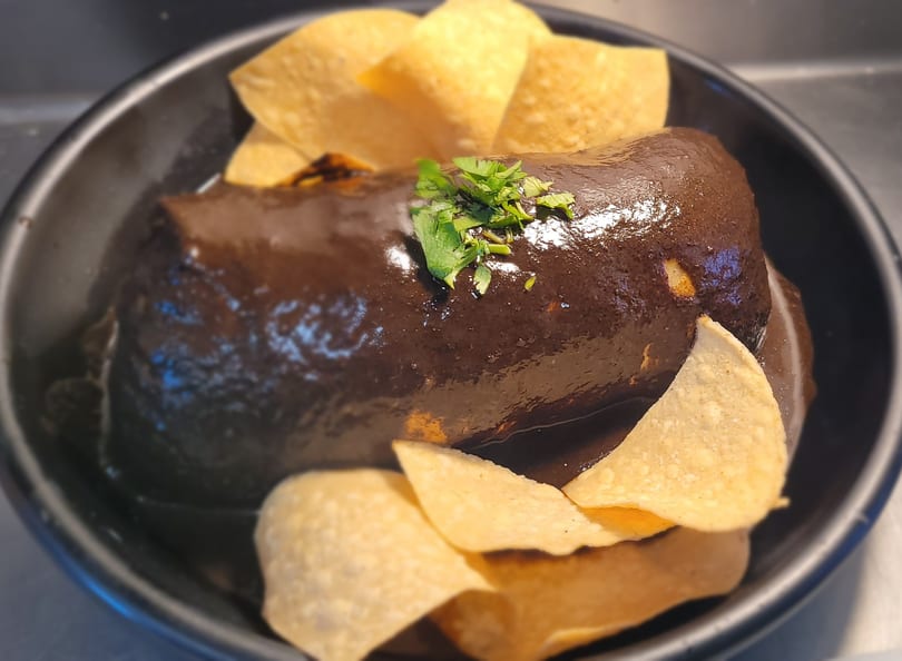 A dark brown mole burrito topped with cilantro and surrounded by tortilla chips in a black bowl.