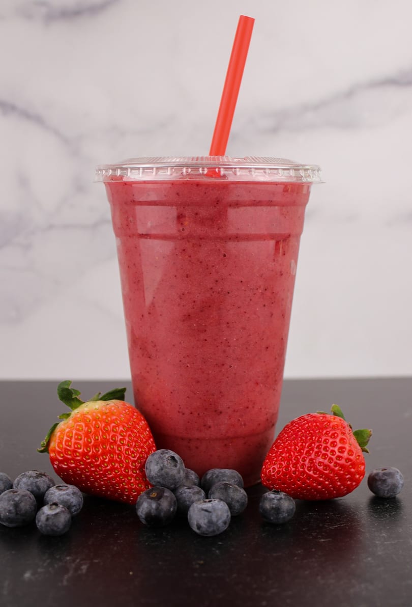 Berry smoothie in a clear cup with a red straw, surrounded by fresh strawberries and blueberries.