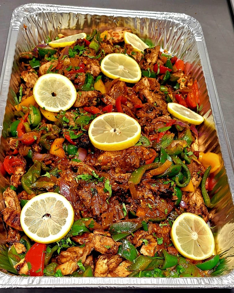 A large foil tray filled with seasoned chicken, colorful bell peppers, onions, and lemon slices.