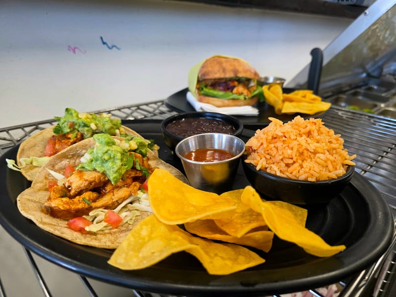 Two chicken tacos with guacamole, rice, black beans, chips, and a burger on a black tray.