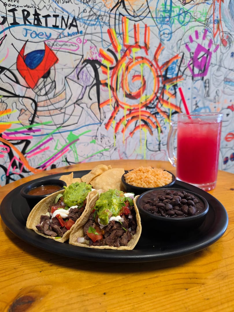 Two tacos with guacamole, rice, beans, chips, salsa, and a red drink on a wooden table.